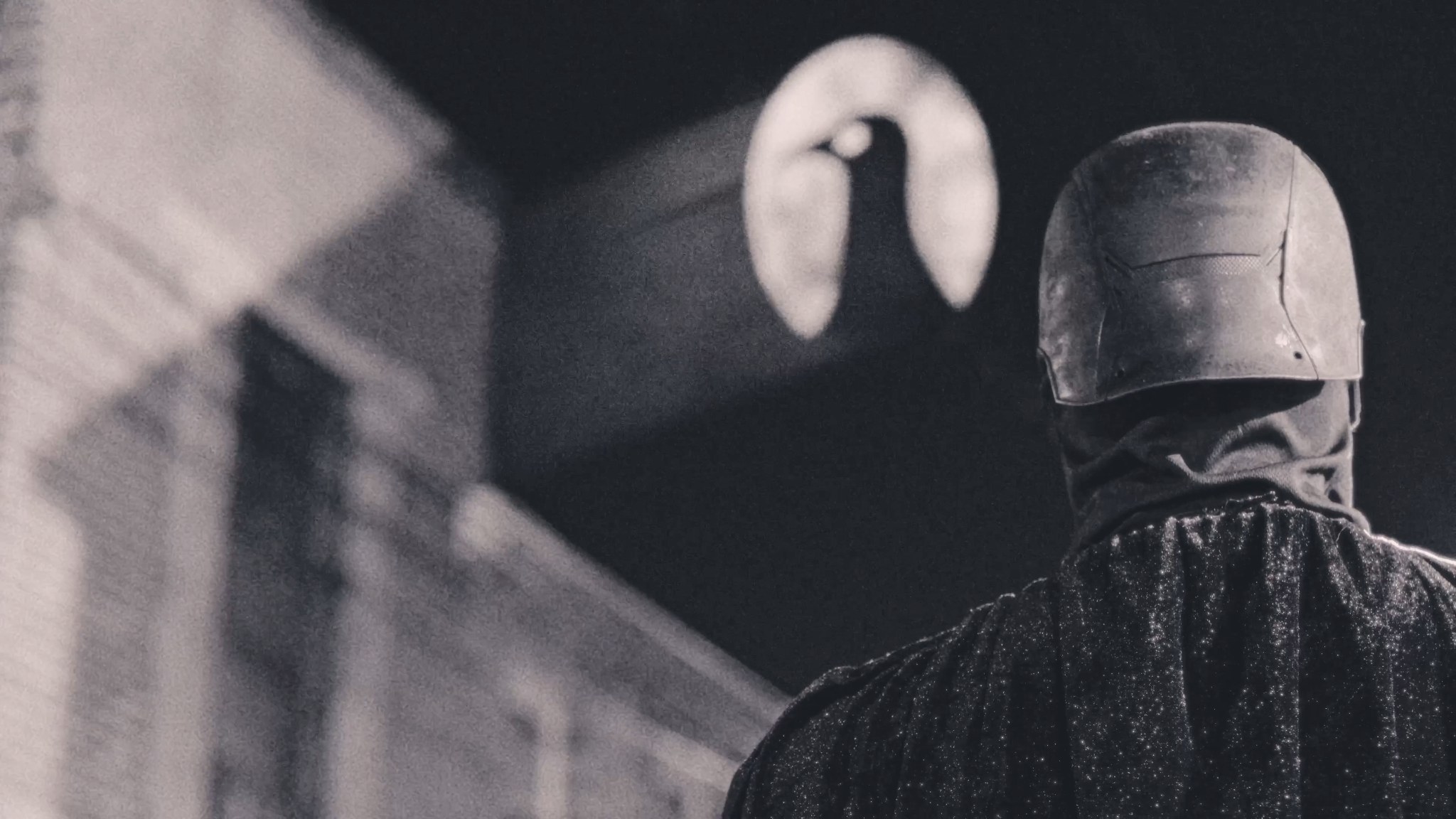 The Raven seen from behind beneath a projected raven symbol on a brick wall.