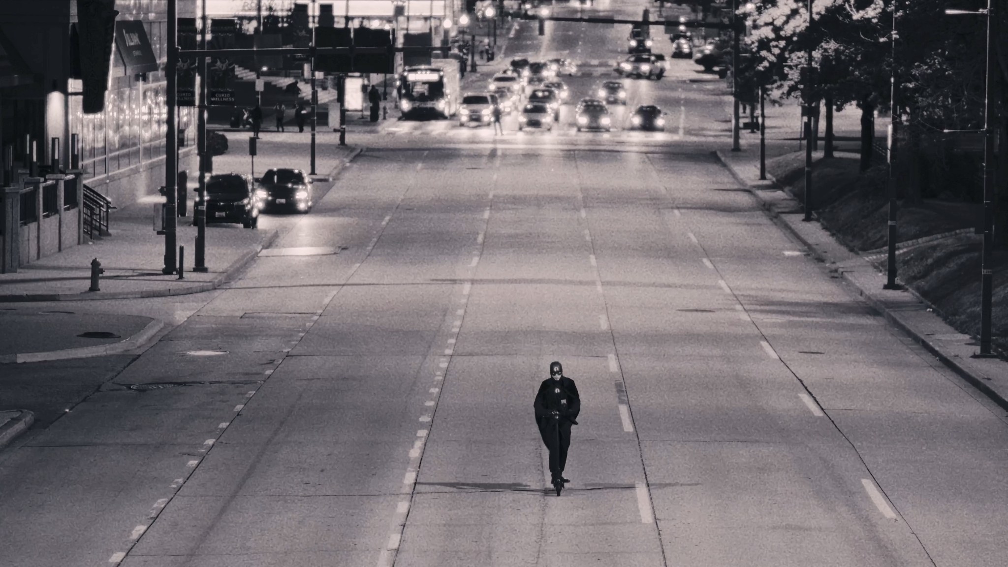 A distant view of the Raven riding a scooter down the center of a broad city street.